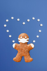 Gingerbread man with protective face mask isolated on blue background with heart created from small sugar snowflakes. Christmas, New year or love concept. Coronavirus winter