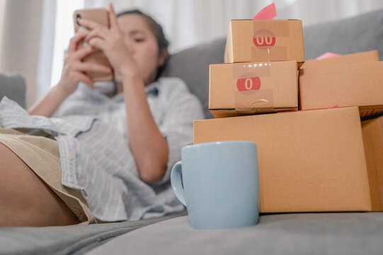 Asian woman business owner working from home with packaging boxes at work She is lying on the sofa and is on the phone to reply to messages from customers who have ordered. Business online concept.