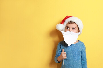 Cute little boy with Santa hat and white beard prop on yellow background, space for text. Christmas celebration
