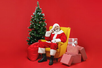 Pensive Santa Claus man in Christmas hat suit coat sit in armchair with fir tree presents gifts put hand on head isolated on red background studio. Happy New Year celebration merry holiday concept.