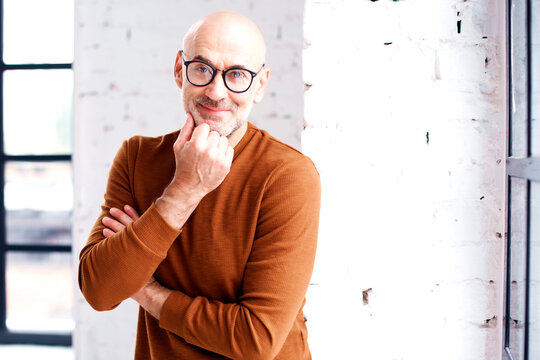 Smiling senior man wearing casual clothes while standing at the window and looking thoughtfully