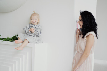 young family, little girl child of European appearance blonde with ponytail hair playing at home clapping her hands. happy brunette mom in beige dress