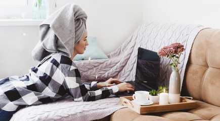 A relaxed woman in a plaid shirt with a towel on her head is working on a laptop doing online...