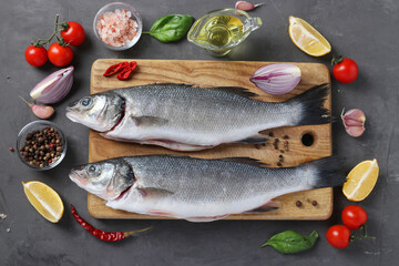 Raw fish seabass with ingredients and seasonings like basil, lemon, salt, pepper, cherry tomatoes and garlic on wooden board on dark background. View from above