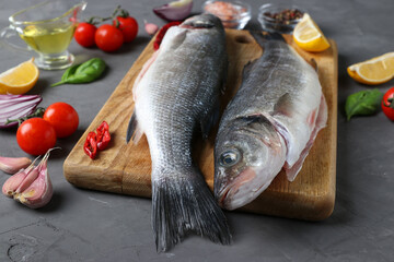 Two raw fish seabass with ingredients and seasonings like basil, lemon, salt, pepper, cherry tomatoes and garlic on wooden board on dark background. Closeup. Horizontal format
