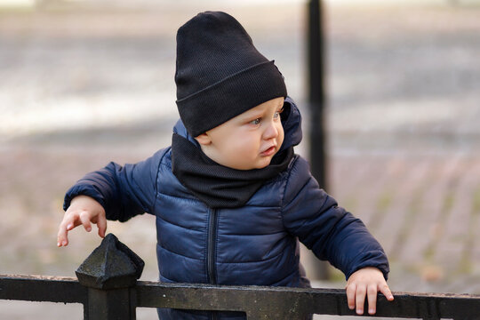 Little Boy In A Black Hat And Jacket. The Child Looks Away With Fear