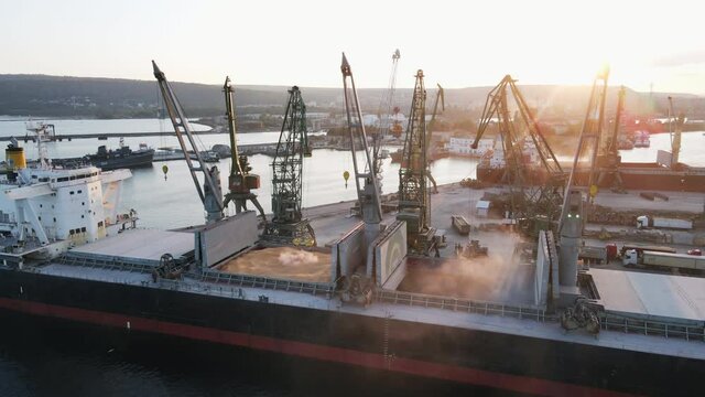Loading of dry cargo ship of wheat by cranes in port for import of transportation on water against sunset aerial view. Export and import of dry cargoes world. Large cargo compartments in hold of ship
