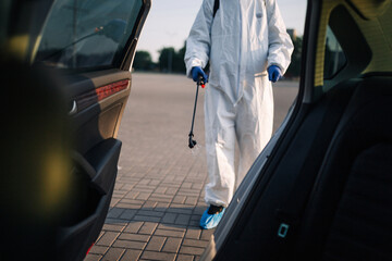 A man wearing protective suit, mask and gloves disinfects the car with a spray to stop spreading of coronavirus disease. Covid-19 pandemic quarantine and cleaning with sanitizer concept.