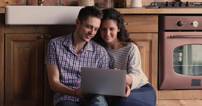 Attractive millennial 30s wife and husband sitting on floor in kitchen spending free time with portable computer, discussing purchase, talking making on-line order use easy comfort internet services