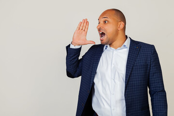 Portrait of a successful young African American businessman guy yelling and shouting loudly to the side with hand over his mouth. Communication concept. Standing on a gray background