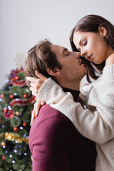 passionate man embracing girlfriend in sweater near christmas tree on blurred background