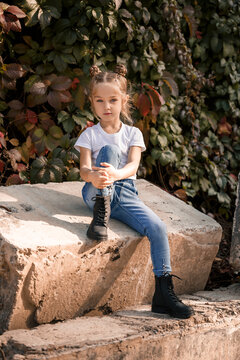 Street photo of a little beautiful girl in jeans and a white t-shirt on a background of concrete slabs and autumn leaves