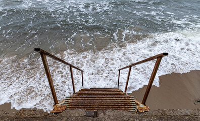 An old rusty metal staircase descending into the raging sea. Abandoned objects on the sea coast.