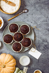 Gluten-free pumpkin chocolate muffins on a gray plate surrounded by ingredients, next to a white card for the inscription. Copy space, flat lei. Healthy baking concept