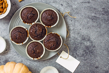 Gluten-free pumpkin chocolate muffins on a gray plate surrounded by ingredients, next to a white card for the inscription. Copy space, flat lay. Healthy baking concept
