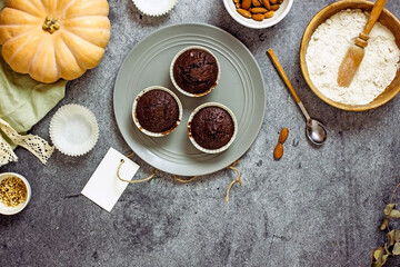 Gluten-free pumpkin chocolate muffins on a gray plate surrounded by ingredients, next to a white card for the inscription. Copy space, flat lay. Healthy baking concept