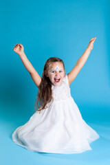 a happy, laughing girl in a white Christmas dress raises her hands in the air, isolated on a blue...