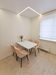 Small kitchen corner with white walls, a dining table and beige armchairs. Vase with roses as an element of interior design on the table in the room. Interior of a modern apartment in white and beige