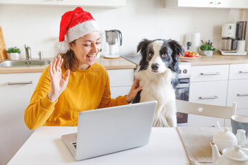 Dog and woman wearing Santa hat waving hand video calling family by webcam. Girl with laptop having virtual meeting chat on holidays sitting on kitchen at home. Happy Christmas and New Year new normal