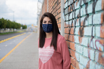 Latin woman, with straight hair, pink sweater, wearing blue masks to protect herself from the coronavirus, in an urban landscape standing next to a wall of red bricks and graffity, behind an unfocused