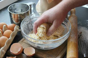 Hands kneading dough. Ball of dough on a rustic wooden background with dusting of flour. Dough with flour near broken egg with yolk in bowl and other utensil, ingredients lies on dark concrete table. 