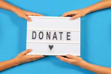 Two person hands holding light box with Donate word text over light blue background. Top view