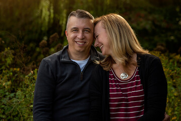 Outdoor close up of sweet middle-age married couple having a romantic moment close together, woman in profile leaning forehead against man who is looking at camera