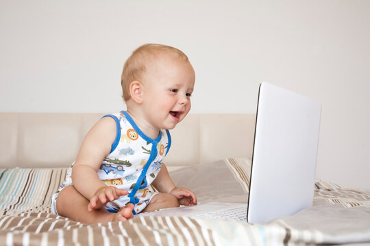 Funny cute child boy sits on a bed in front of an open computer laptop and emotionally looks at the screen.