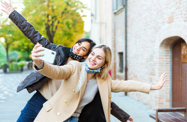 Fototapeta premium Young people taking selfie on city street wearing face protective masks.Coronavirus prevention, travel safe, technology trends and autumn vacation concept. couple of funny women during new normal