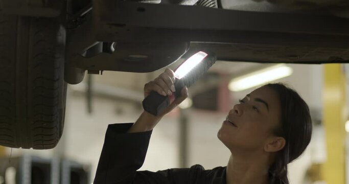 Female car mechanic examining vehicle using flashlight in automotive vehicle repair workshop