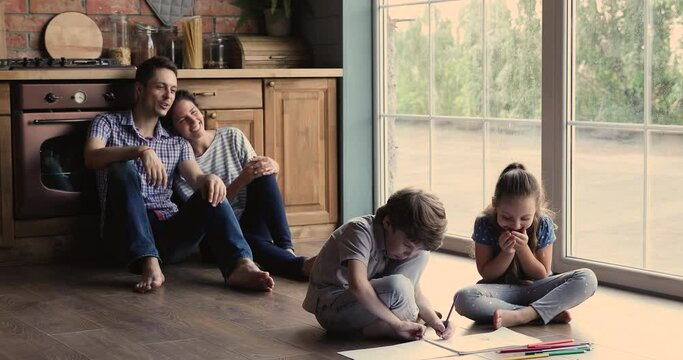 Full family sit on warm floor with underfloor heat system in kitchen, little children draw with colored pencils on album while parents looking at kids, spend weekend free time at modern house concept