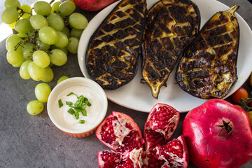 Roasted eggplants on white plate, pomegranates and grapes on a table. Mediterranean style food. 