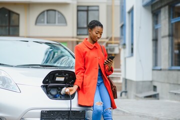 Charging electro car at the electric gas station. African American girl standing by the car.