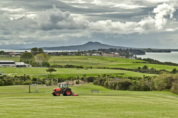 AUCKLAND, NEW ZEALAND - Mar 28, 2019: Lawn mower red tractor working in Macleans park with Hauraki Gulf islands in background © Bearhunter/Wirestock