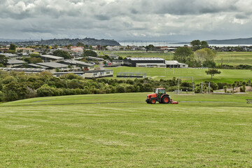 AUCKLAND, NEW ZEALAND - Mar 28, 2019: Lawn mower red tractor working in Macleans park with Hauraki Gulf islands in background © Bearhunter/Wirestock