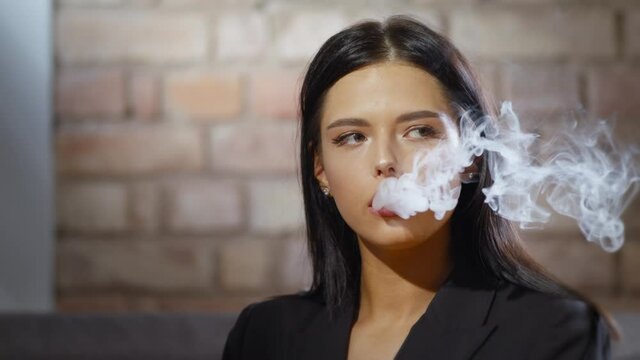 Dark-haired young woman smoking electronic cigarette on brick wall background