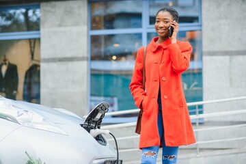 Fototapeta premium Using smartphone while waiting. African American girl on the electric cars charge station at daytime. Brand new vehicle.