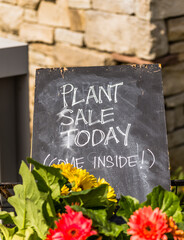 A plant sale today sign sits among some flowers outside a garden center inviting people inside.