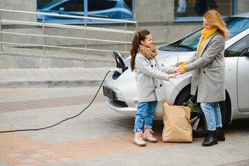 mother with daughter charging electro car at the electric gas station and speak on mobile phone.