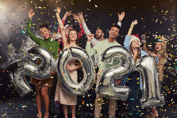 Happy New Year. Group of young excited multiracial friends holding silver foil balloons in form of numbers 2021 and raising hands up, confetti falling in the air