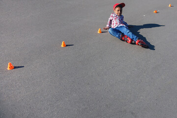 Little african american boy on rollers skates lies at asphalt road among training cones . Roller...