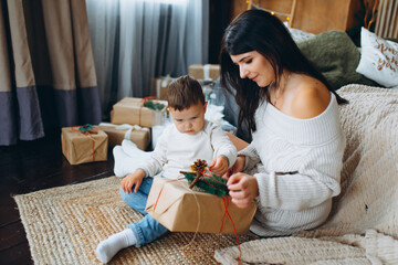 beautiful brown haired mom and lovely boy son sitting on the floor and opening gifts in a box on the background of the Christmas tree