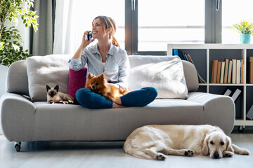 Pretty young woman talking with mobile phone while sitting in couch with her dogs and cat at home.