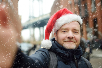 Local man in a Santa Claus hat takes selfie on street near the Manhattan Bridge in New York City on Christmas Eve during a snowfall. Winter Xmas holidays in NYC.