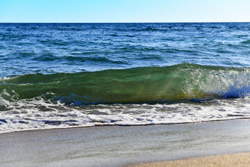 Sea coast. A large greenish-blue wave rose over the sandy shore. White foam rolls off the beach.