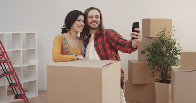 Happy couple taking selfie among cardboard boxes with things in new apartment. Handsome caucasian guy holding smartphone. People grimace at camera.