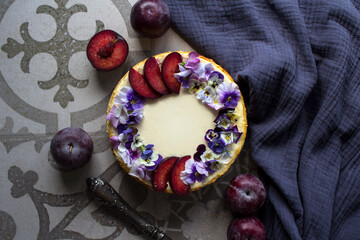 Cheesecake decorated with red plums and wild pansy flowers. Top view photo of classic dessert. 
