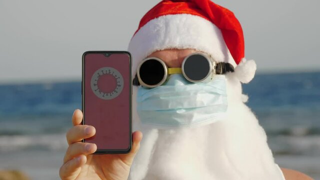 funny santa claus, in a protective mask. Santa, in sunglasses, holds smartphone with COVID-19 icon, text - quarantine, on screen, on beach by the sea. santa claus warns of coronavirus danger
