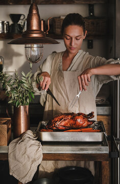 Young woman in apron carving whole oven roasted duck for Thanksgiving holiday or Christmas party celebration in kitchen interior, Traditional Autumn or Winter holiday comfort food concept
