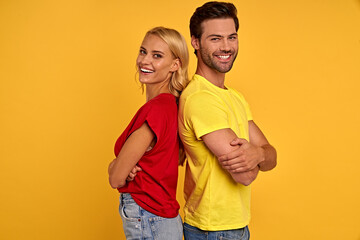 Side view of smiling young couple in red and yellow t-shirts isolated on yellow background. People lifestyle concept. Mock up copy space. Standing back to back holding hands crossed looking camera.